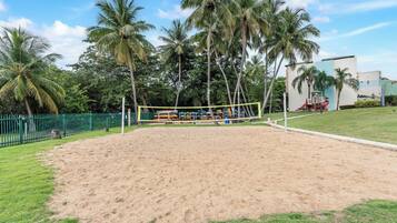 A sandy volleyball court with a net, surrounded by palm trees and a green fence.