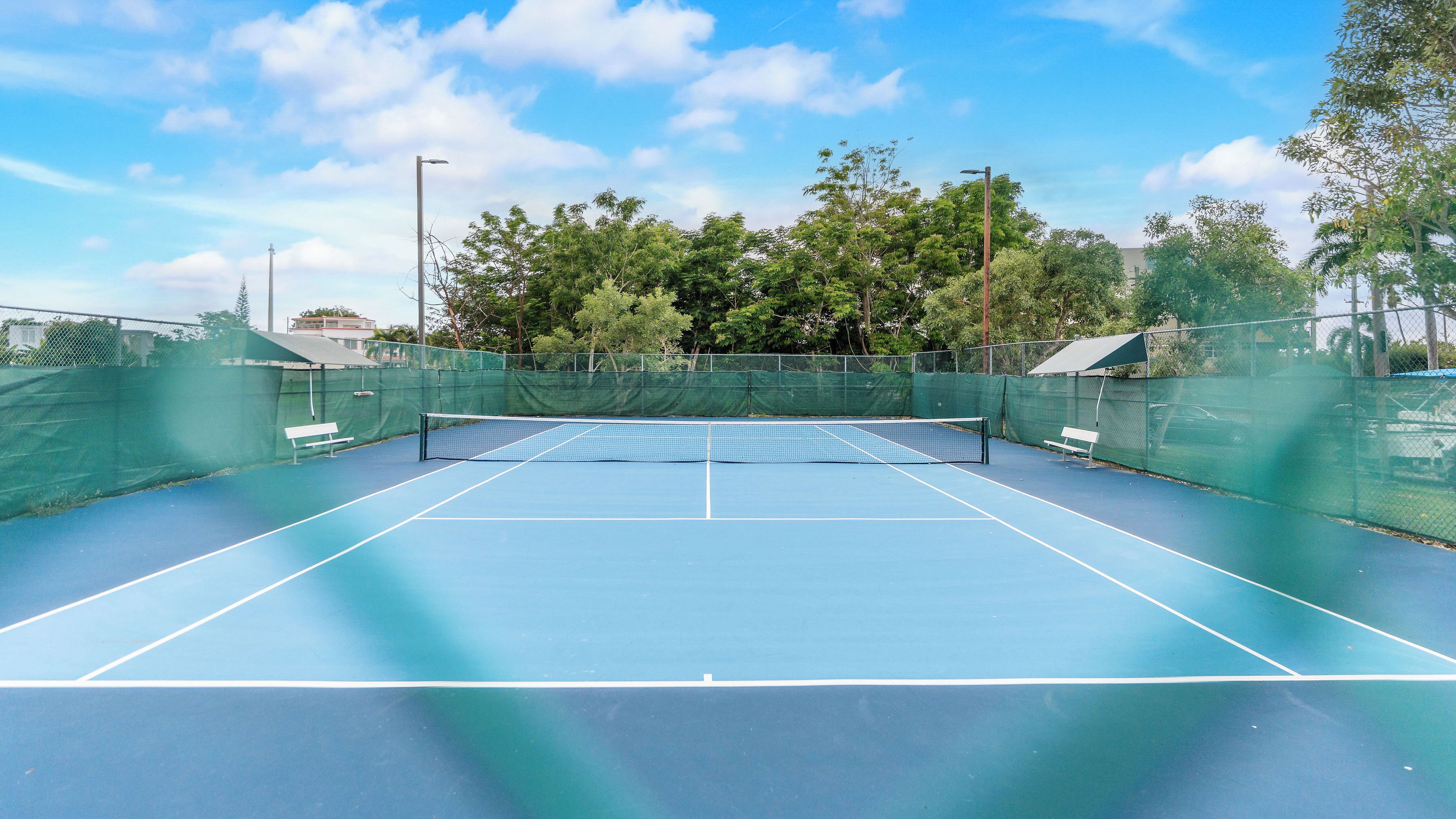 An outdoor tennis court with a net, benches, and a green fence.