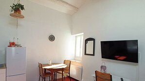Une cuisine compacte équipée d’un réfrigérateur, d’une table à manger avec des chaises, d’une plante suspendue, d’une horloge murale et d’un téléviseur à écran plat fixé au mur.