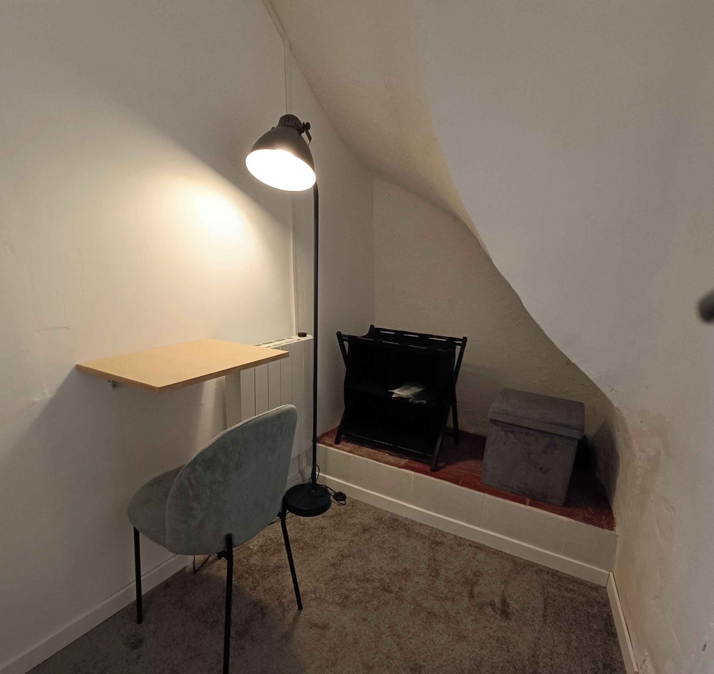 Una habitación pequeña y con poca luz, con un escritorio, una silla y una lámpara de pie.