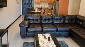 A living room with a black leather sofa, a coffee table with remote controls, and a kitchen in the background. - 2-bedroom condo in delightful Kingston with cool AC (Kingston)