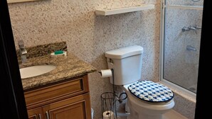 A bathroom with a granite countertop, wooden cabinets, a toilet with a patterned seat, a shower area, and a towel rack. - 2-bedroom condo in delightful Kingston with cool AC (Kingston)