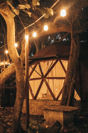 Una casa en el árbol con un muro de piedra y estructura de madera, iluminada con luces de adorno.