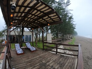 Una terraza de madera con dos tumbonas, un espacio de descanso cubierto y vista a la playa.