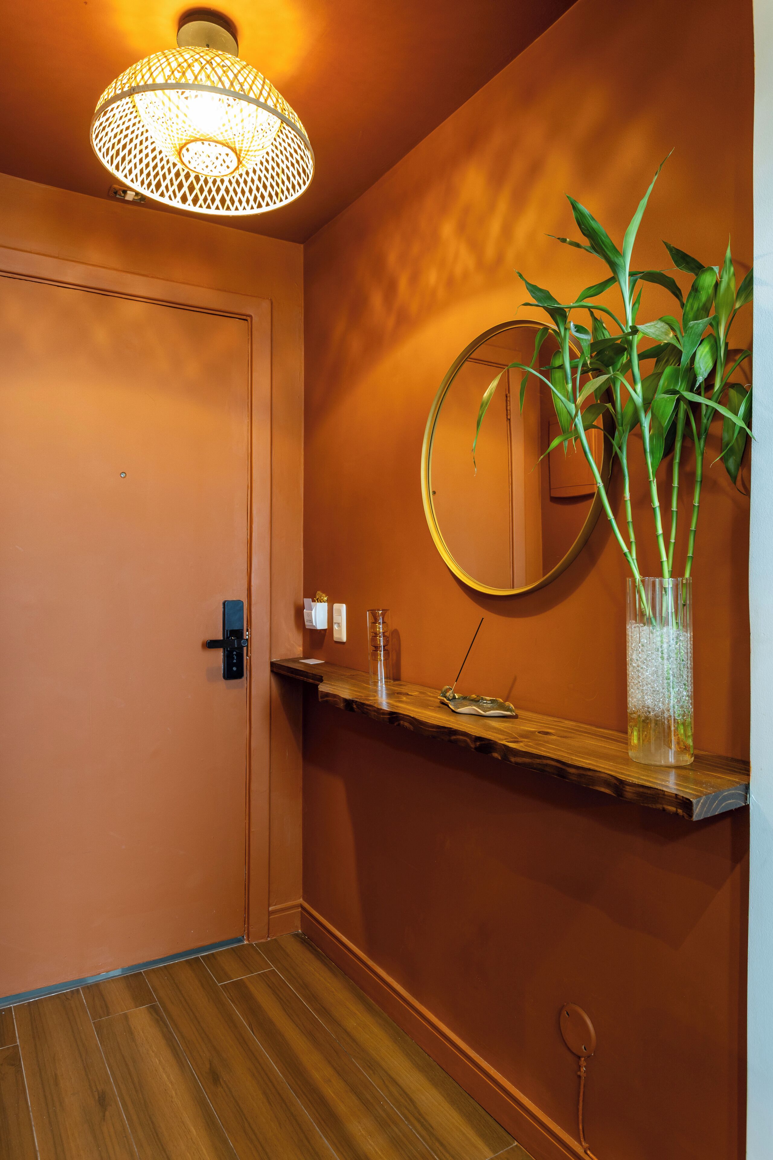 A hallway with a wooden floor, a door, a mirror, a shelf with a plant, and a hanging light fixture.