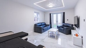 Un salon moderne avec un plafond blanc, deux canapés en cuir noir, une table basse blanche et un téléviseur fixé au mur.