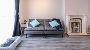 Ett vardagsrum med en grå soffa, två mönstrade kuddar, ett litet sidobord med en kaffekanna och en öppen spis med en dekorativ hylla ovanför.