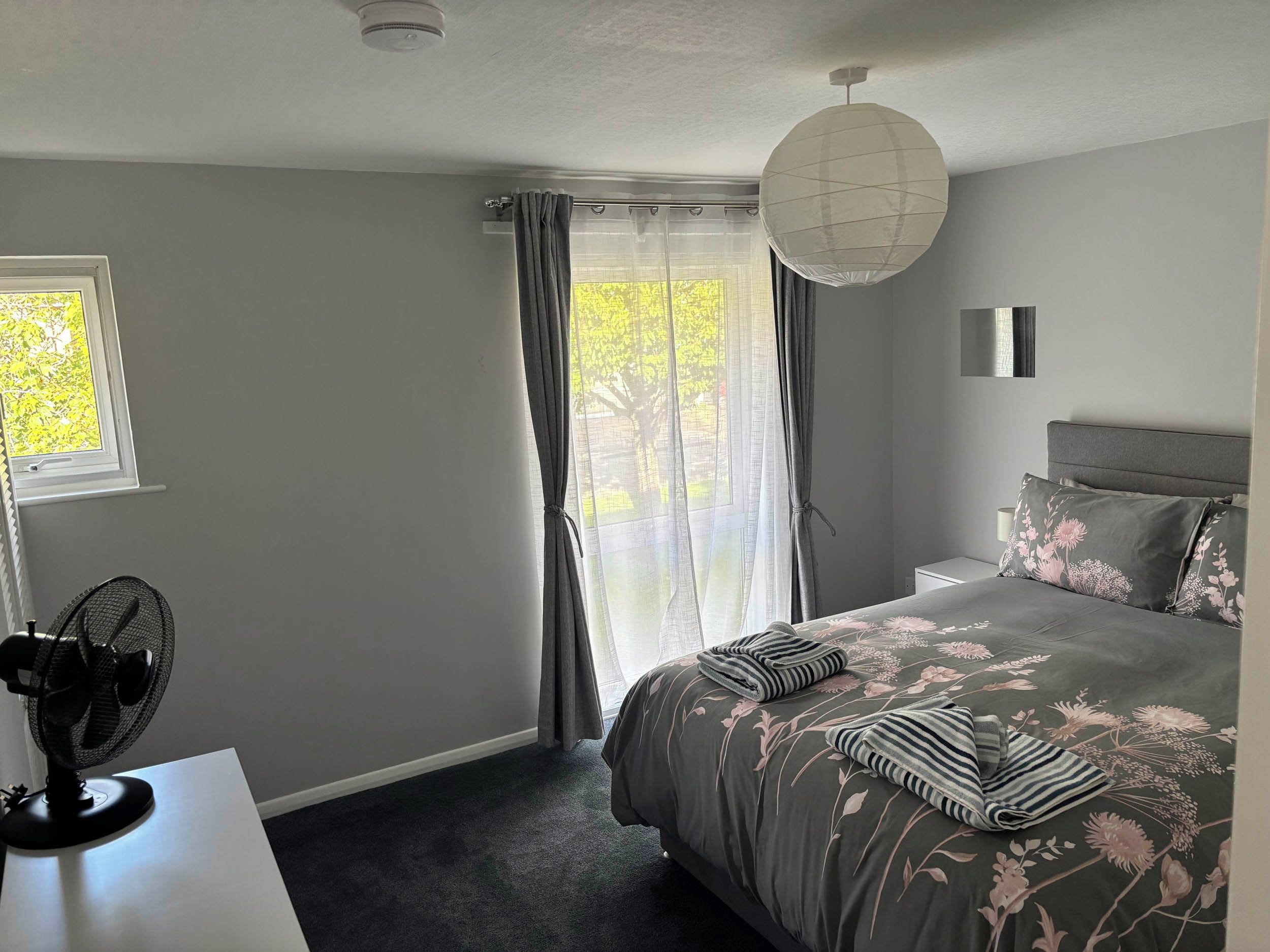 A bedroom with a bed, a bedside table, a fan, and a window with curtains.