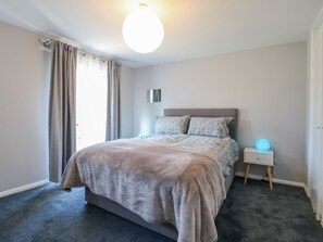 A bedroom with a large bed, a bedside table with a lamp, and a window with curtains. - Sea Rose Broadstairs (Broadstairs)