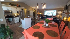 Eine moderne Küche mit Edelstahlgeräten, ein Essbereich mit einem roten Tisch und gewobenen Tischsets sowie ein Wohnzimmer mit Sofa und Couchtisch.