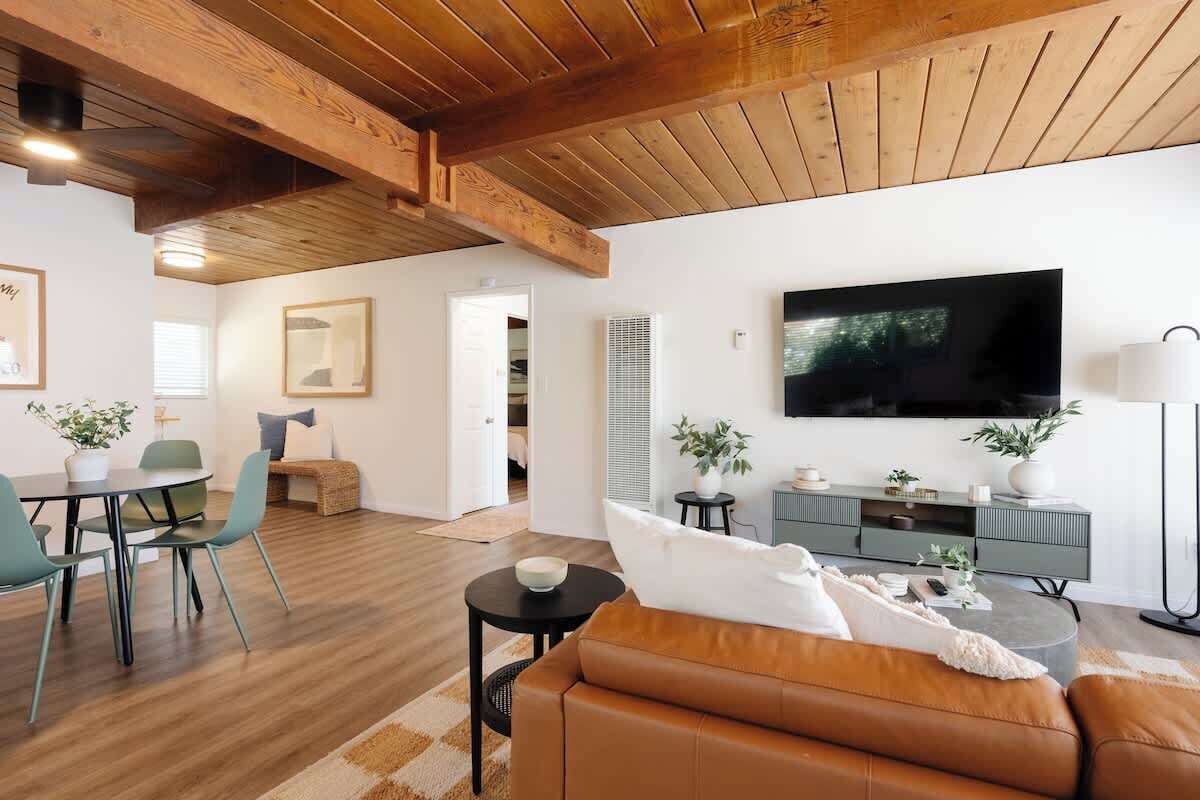 A modern living room with a wooden ceiling, a flat-screen TV, a sofa, a dining area with chairs, and a small table.