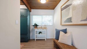 Ein Zimmer mit Holzbalkendecke, einer weißen Wand, einem Fenster mit Jalousien, einem weißen Regal mit Pflanzen, einer Korbsitzbank mit Kissen und einem gerahmten abstrakten Gemälde.