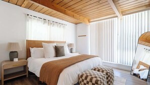 Ein Schlafzimmer mit Holzbalkendecke, einem großen Bett mit weißer Bettwäsche, einem karierten Hocker und einem Nachttisch mit Lampe.