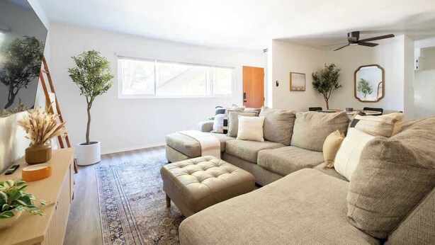 Amplia sala de estar con un sofá seccional grande, una mesa de centro y una zona de comedor con una mesa y sillas de madera. Se aprecia una ventana grande que proporciona luz natural, y un ventilador de techo.