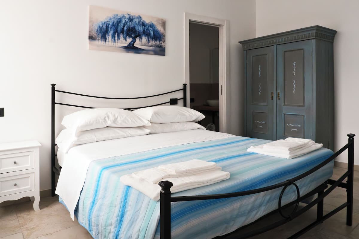 Ein ordentlich bezogenes Bett mit weißen Bettwäsche und einer blau-weiß gestreiften Tagesdecke, ein weißer Nachttisch mit Schubladen sowie ein blauer Kleiderschrank mit dekorativen Elementen.