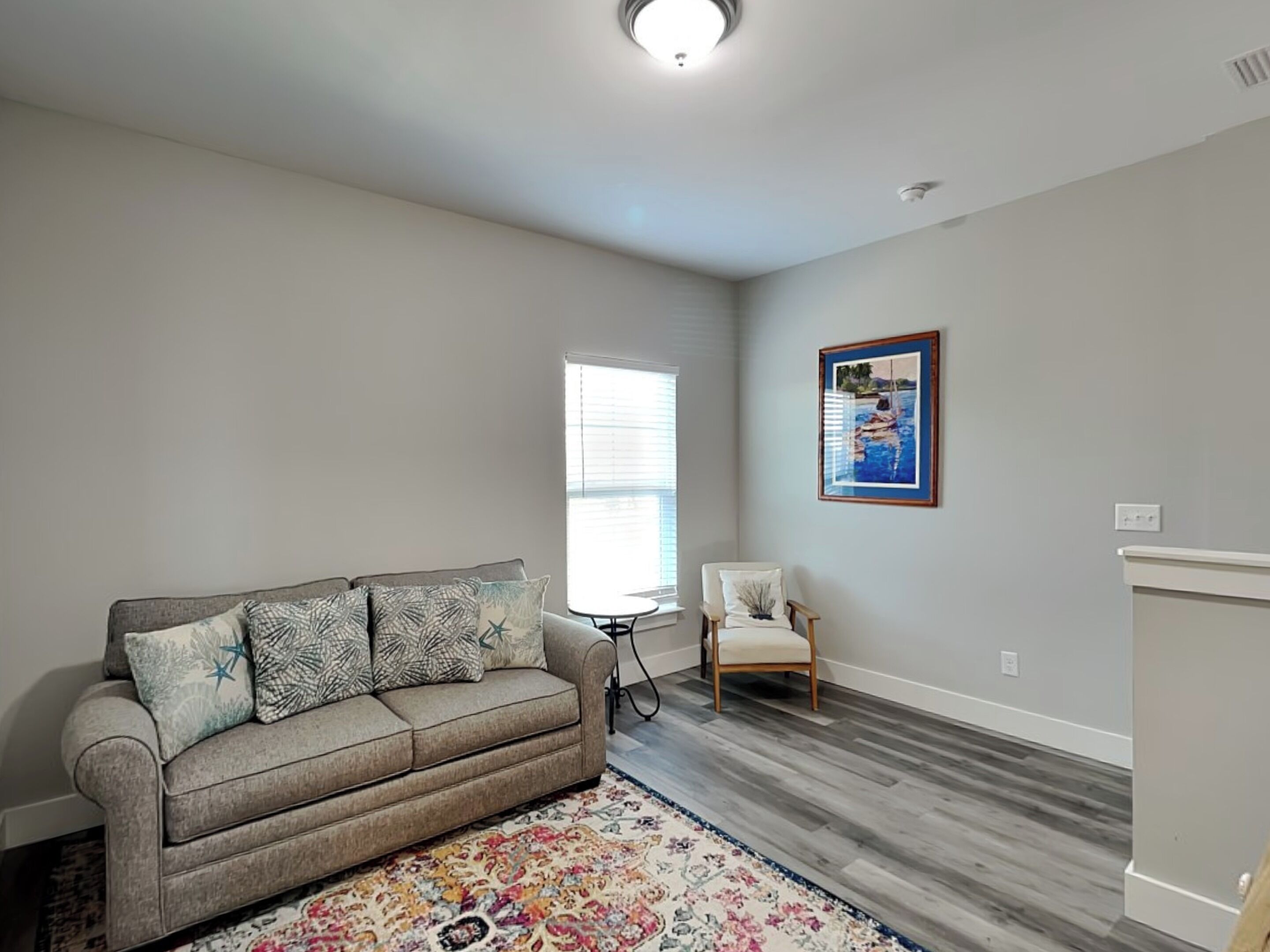 A living room with a grey sofa, a patterned rug, a wooden chair, and a framed picture on the wall.