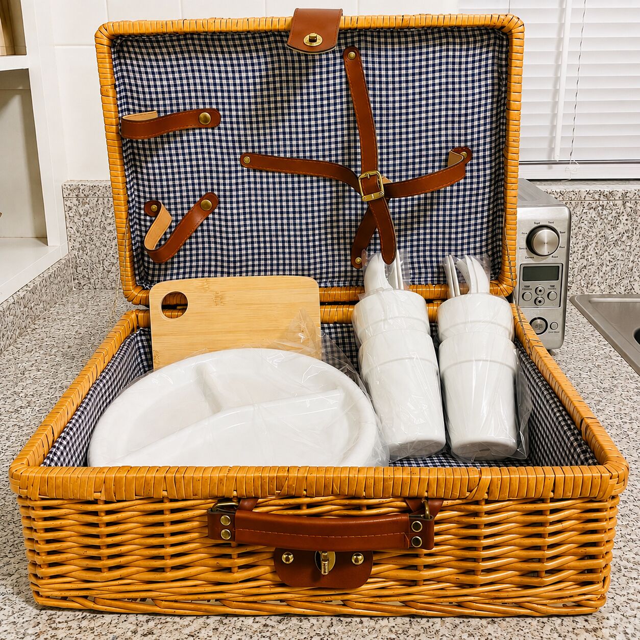 A wicker basket with a checkered interior, containing a wooden cutting board, two white plastic containers, and a white plate.