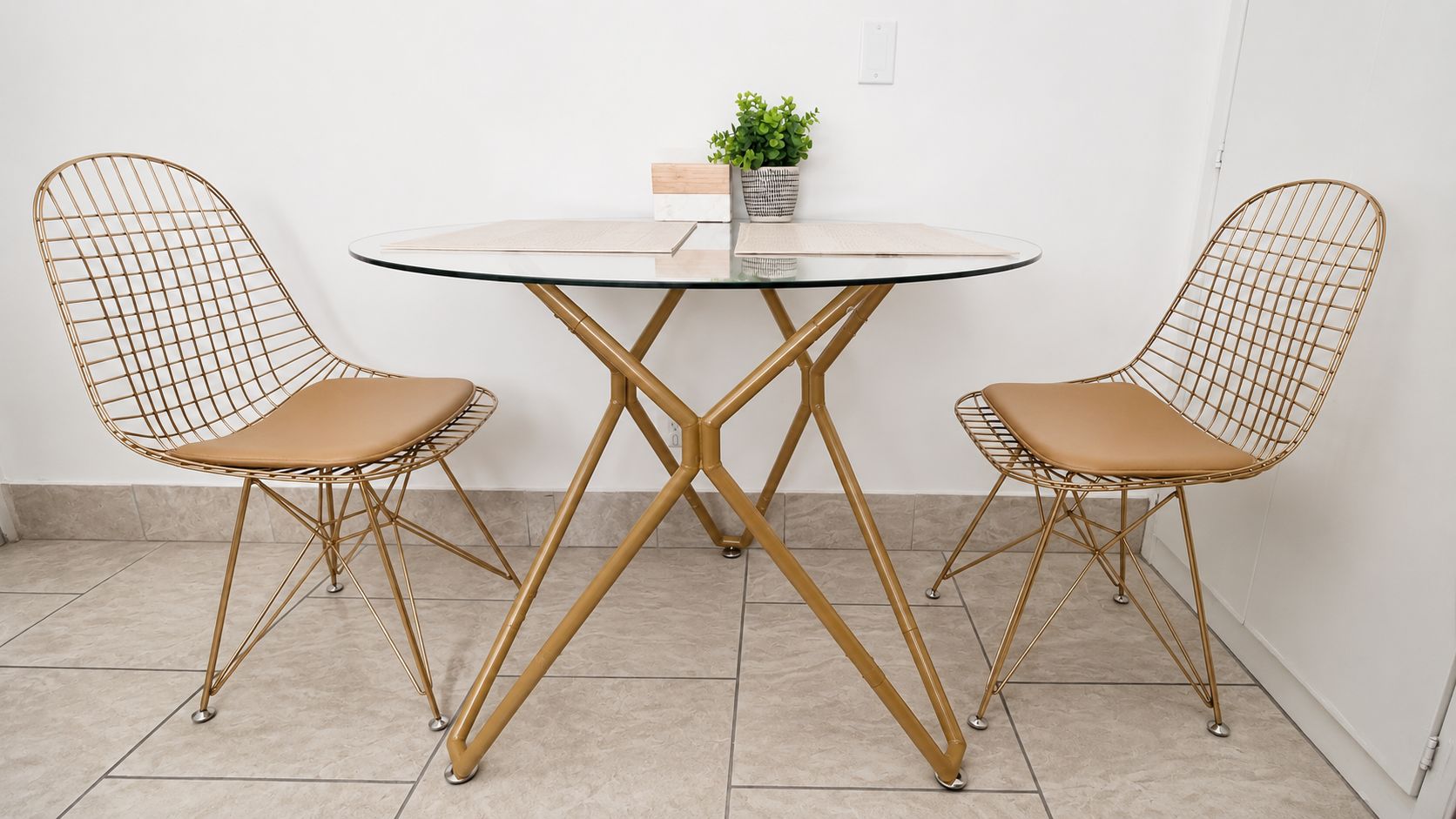 A dining area with two wireframe chairs and a glass table with a central support structure.