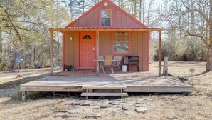 Une petite cabane en bois avec une porte rouge, une véranda avec deux chaises et un barbecue à l’extérieur.