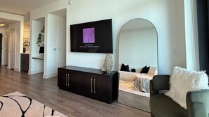 Een moderne woonkamer met een flatscreen televisie aan de muur, een donker tv-meubel, een bank en een grote spiegel.