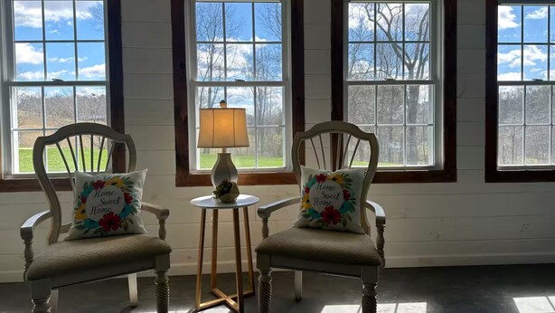 Quarto com duas cadeiras de madeira, cada uma com uma almofada floral, uma mesa pequena com abajur e janelas amplas com vista para árvores e céu.