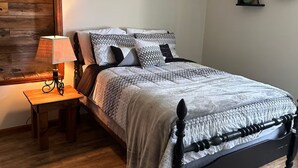 Quarto com cama, mesa de cabeceira de madeira, abajur, ventoinha de teto e uma parede com prateleiras.