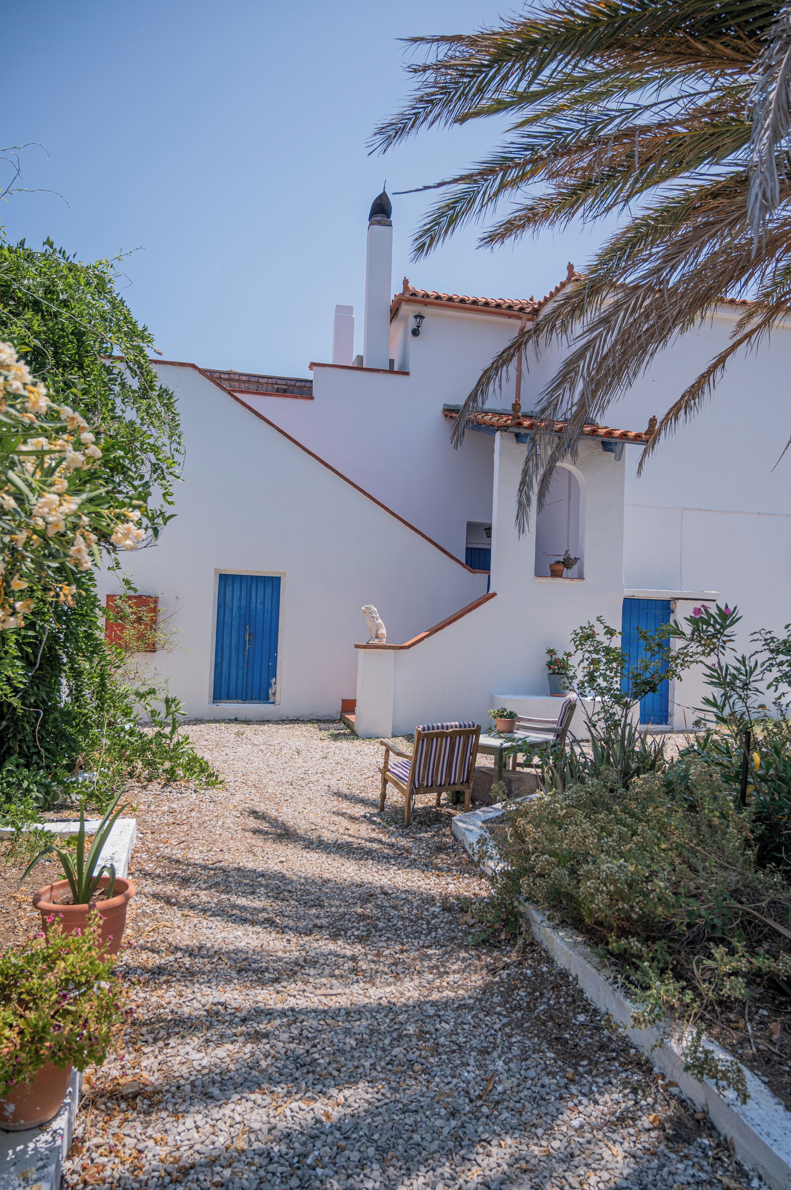 A white building with a blue door, a small balcony, and a gravel path leading to a table and chairs.