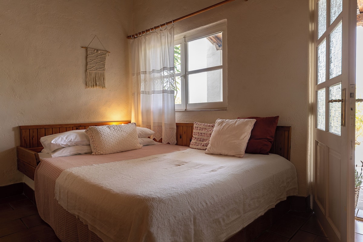 A bedroom with a large bed, a bedside table, a lamp, a window with curtains, and a wall-mounted decoration.