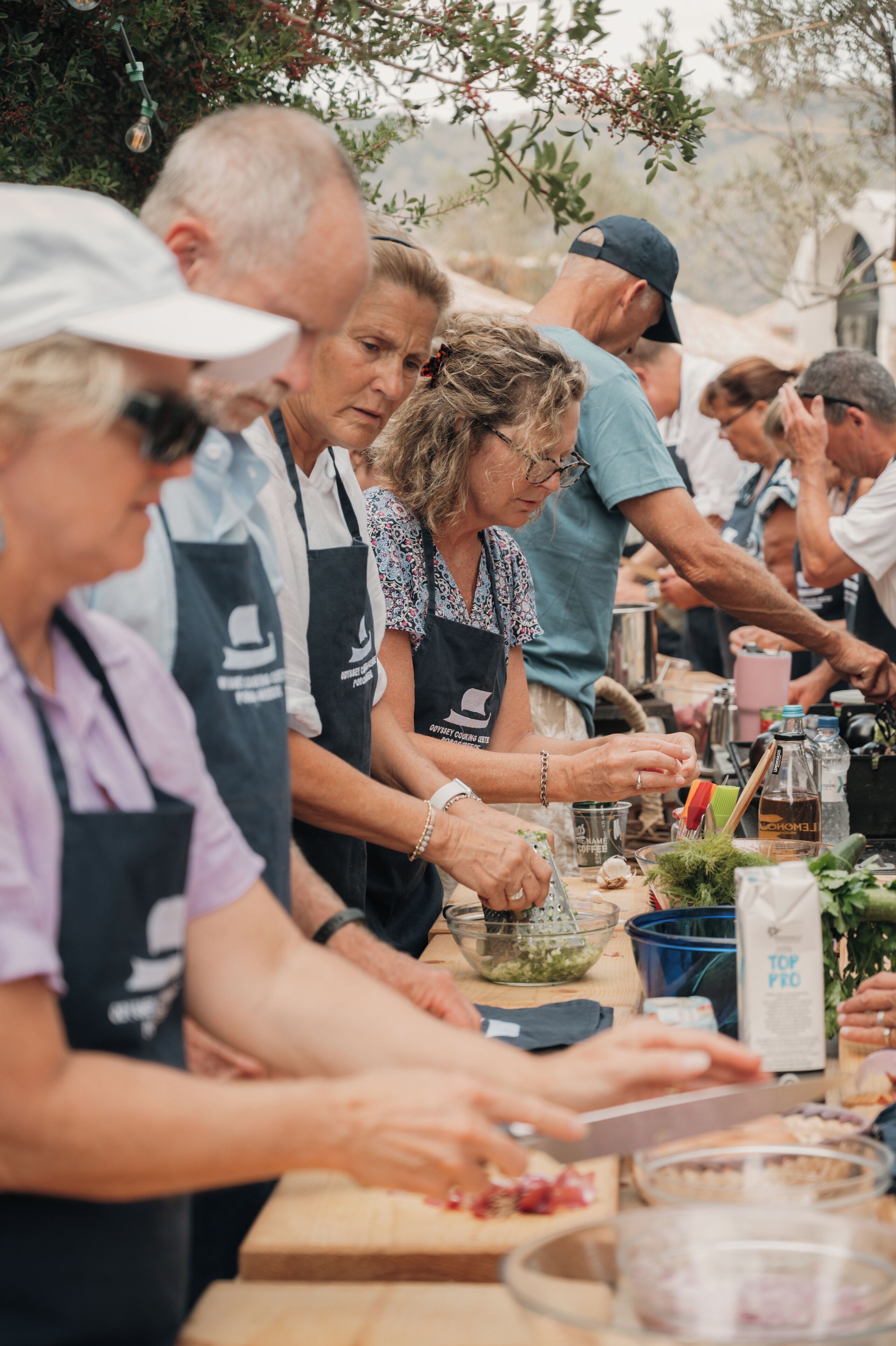 Eine Gruppe von Menschen bereitet im Freien Essen zu, sie tragen Schürzen, und ein Tisch ist voll mit Zutaten und Kochgeschirr.