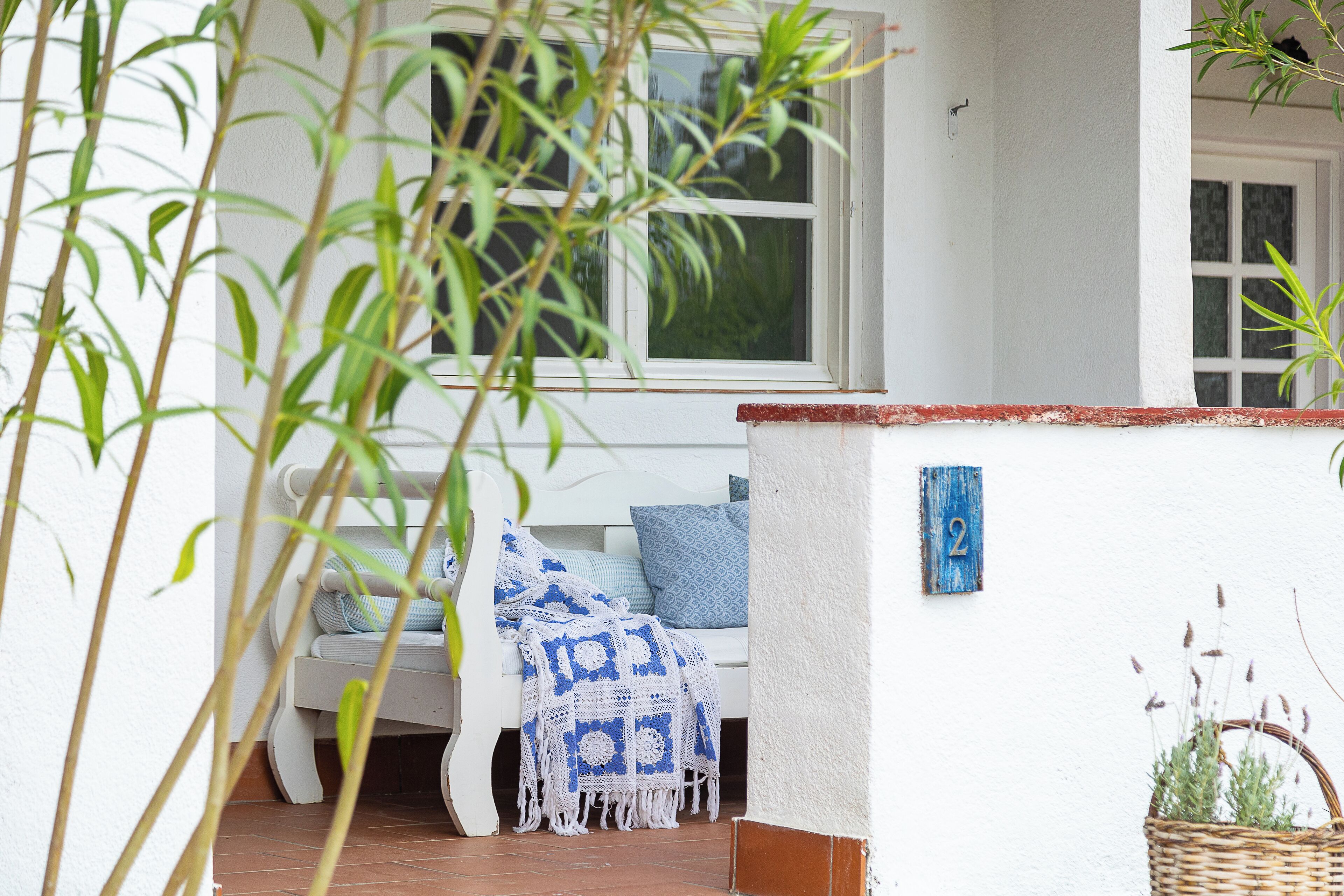 A white bench with a blue and white patterned blanket, a wicker basket with plants, and a number '2' sign on a white wall.