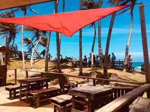 Ein Sitzbereich am Strand mit Holztischen und -stühlen, einem roten Sonnensegel und Palmen.