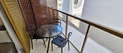 A balcony with a round glass table and two chairs, a metal railing, and a view of adjacent buildings.