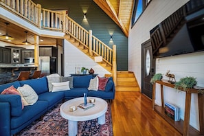 A spacious living room with a blue sectional sofa, a coffee table, and a staircase leading to a loft area. - NEW! Stunning 4BR Cabin w/Hot Tub, Fire Pit, Pool Table, Arcades & Pool Access (Sevierville)