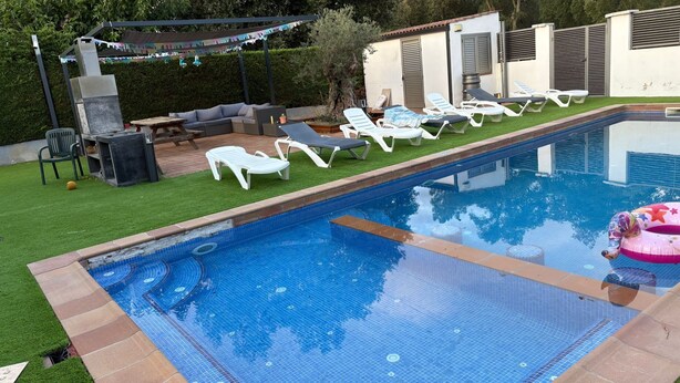 En swimmingpool med flisekant, omgivet af en græsplæne med liggestole og grillområde.