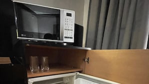 A compact kitchen with a microwave on the countertop, a small refrigerator with a single door, and a wooden shelf above the fridge. - FARIA LIMA HOTEL COM VARANDA GARDEN  (São Paulo)