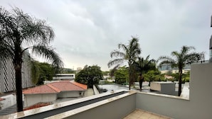 A rooftop terrace with tiled flooring, palm trees, and a view of a building with a red-tiled roof. - FARIA LIMA HOTEL COM VARANDA GARDEN  (São Paulo)