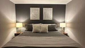 Ein ordentlich bezogenes Bett mit dunklem Kopfteil, zwei Nachttischlampen und abstrakten Wandbildern.