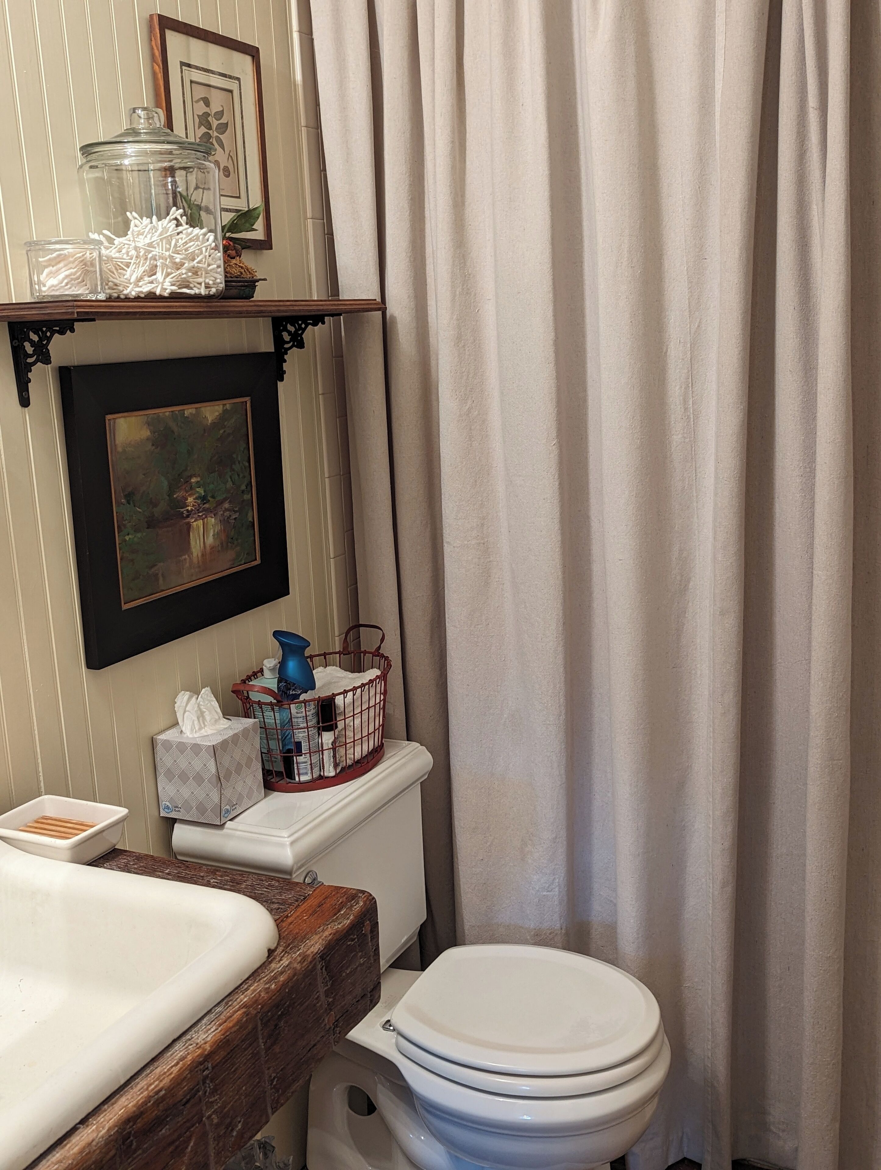 A bathroom with a white toilet, a wooden vanity, a framed painting, a glass jar, and a basket on the wall.
