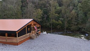 A cabin with a wooden exterior and a red roof, situated on a gravel area with a view of a forest and a stream. - Amazing Waterfalls & Swim Hole at Dale Hollow Lake (albany)