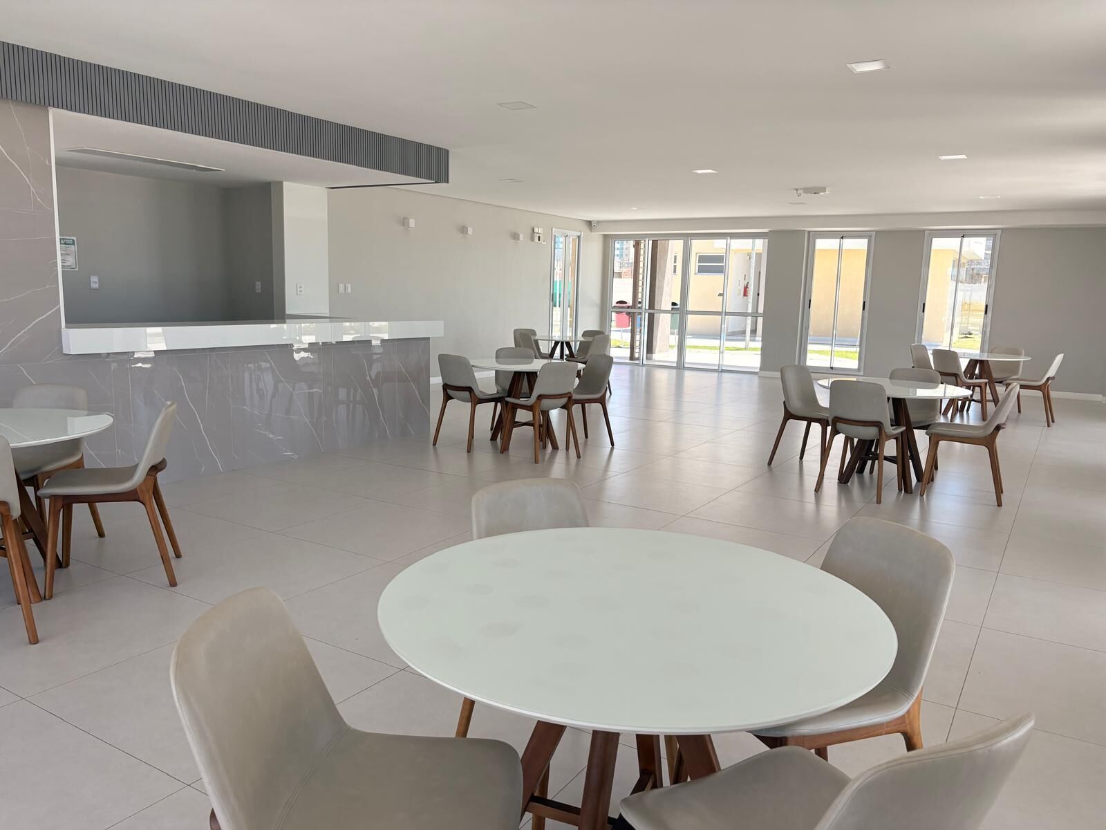 A spacious indoor area with multiple round tables and chairs, a kitchen counter, and large windows allowing natural light.