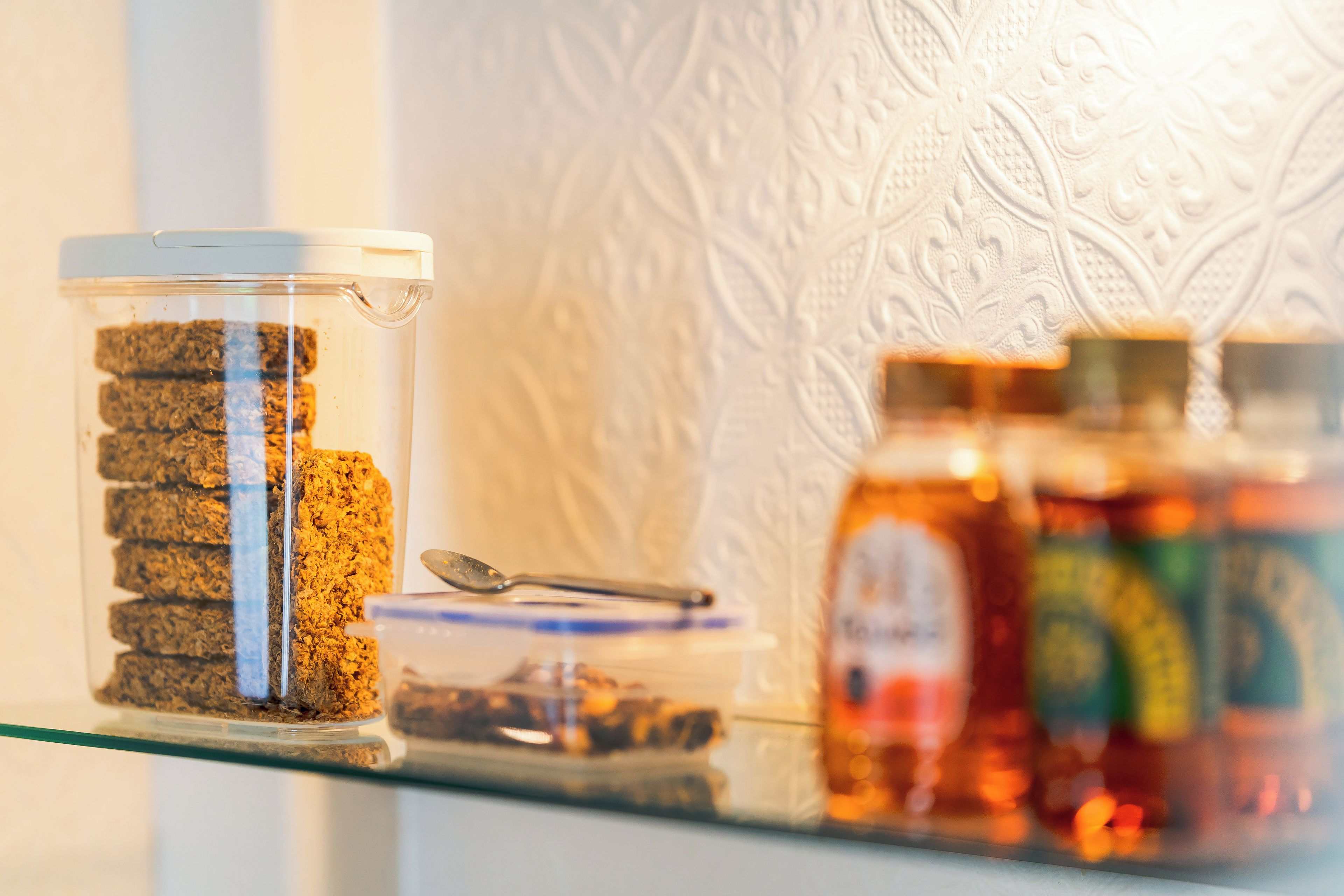 A glass jar with a spoon on a shelf, next to a patterned wall.