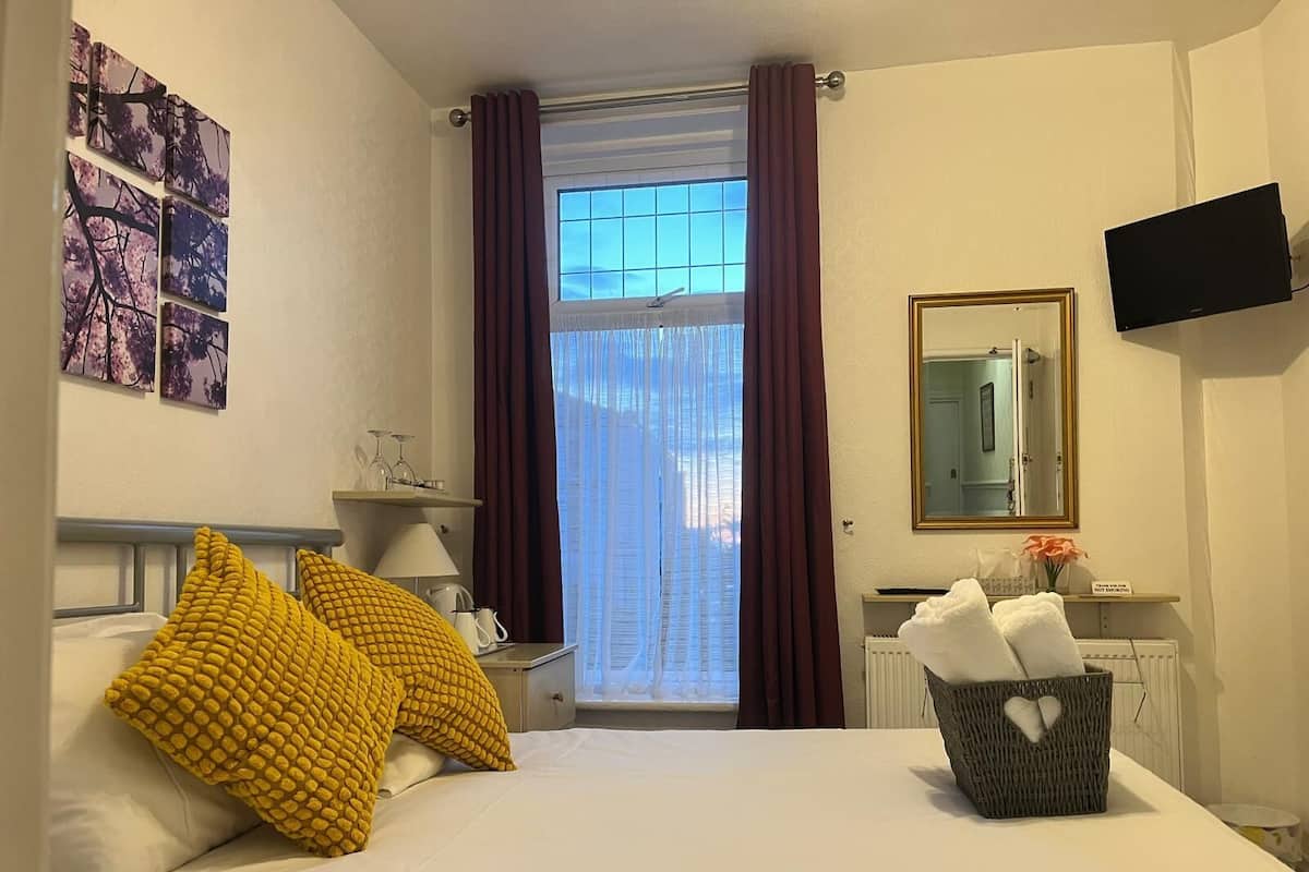 Hotellihuone, jossa on sänky, yöpöytä lampun kanssa, televisio, ikkuna verhoilla ja taideteos seinällä.