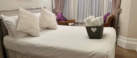 A neatly made bed with white bedding and pillows, a wicker basket with towels, and a window with curtains.