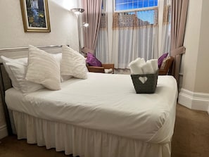 Un lit bien fait, avec du linge de lit et des oreillers blancs, un panier en osier rempli de serviettes et une fenêtre avec des rideaux.