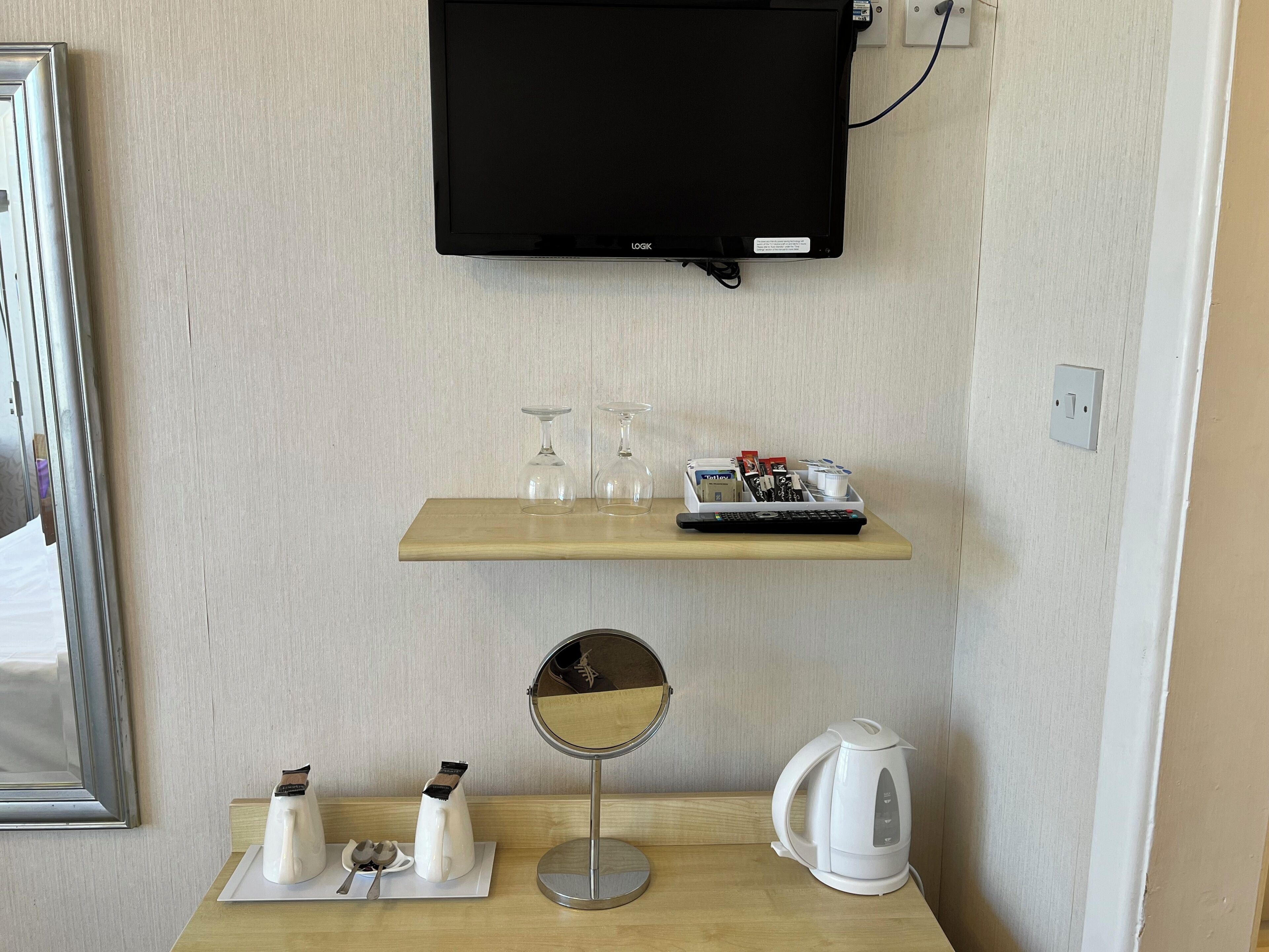 A hotel room with a mounted TV, a shelf with glasses and a remote, a mirror, and a kettle on the table.