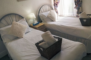 Ett hotellrum med två sängar, var och en med en kudde och en näsdukshållare. Vid ena sängen står ett nattduksbord med en lampa och en korg.