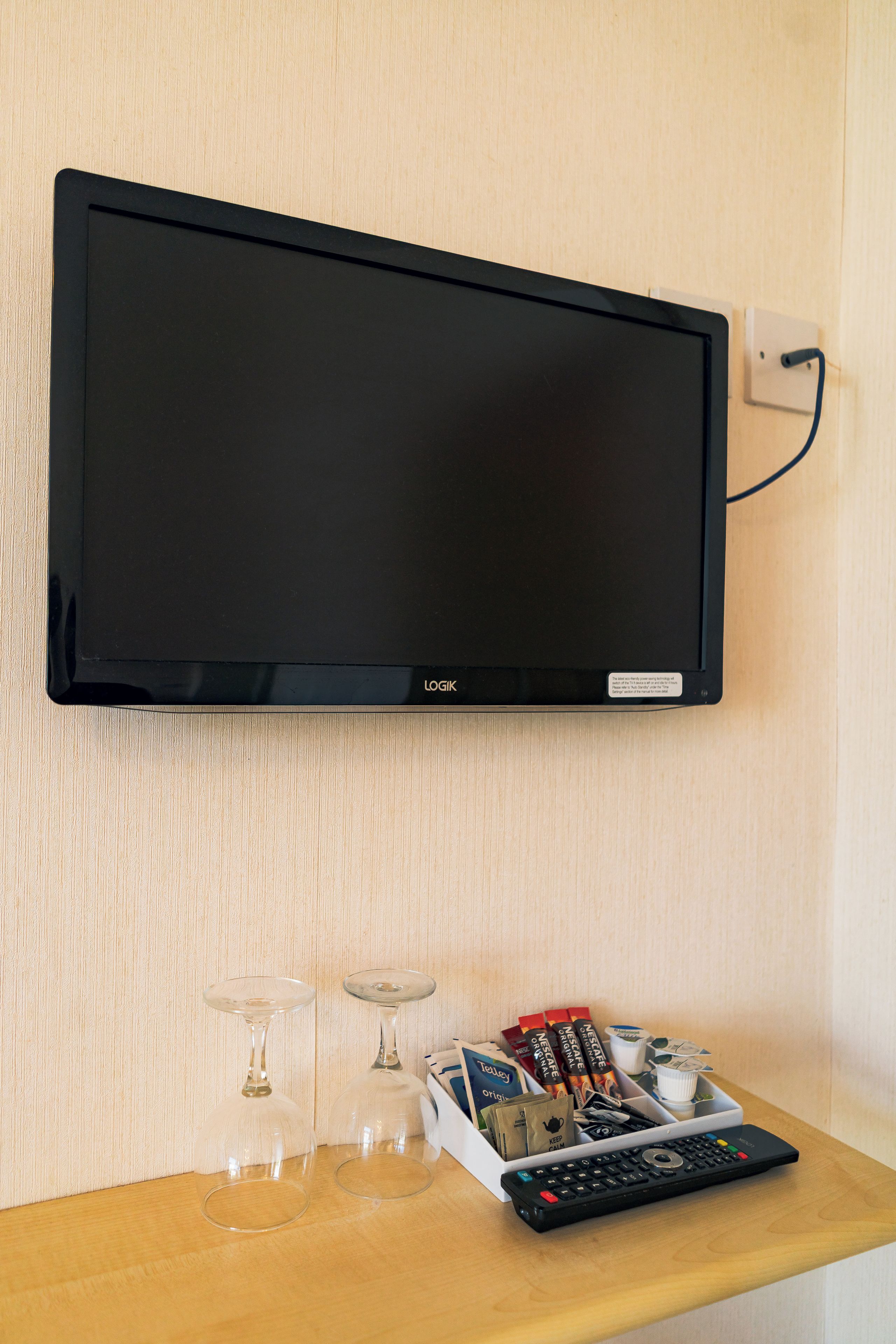 A flat-screen TV mounted on a wall, a wooden table with a remote control, two wine glasses, and a box of Nescafe Original coffee.