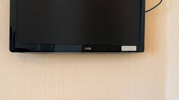A flat-screen TV mounted on a wall, a wooden table with a remote control, two wine glasses, and a box of Nescafe Original coffee.