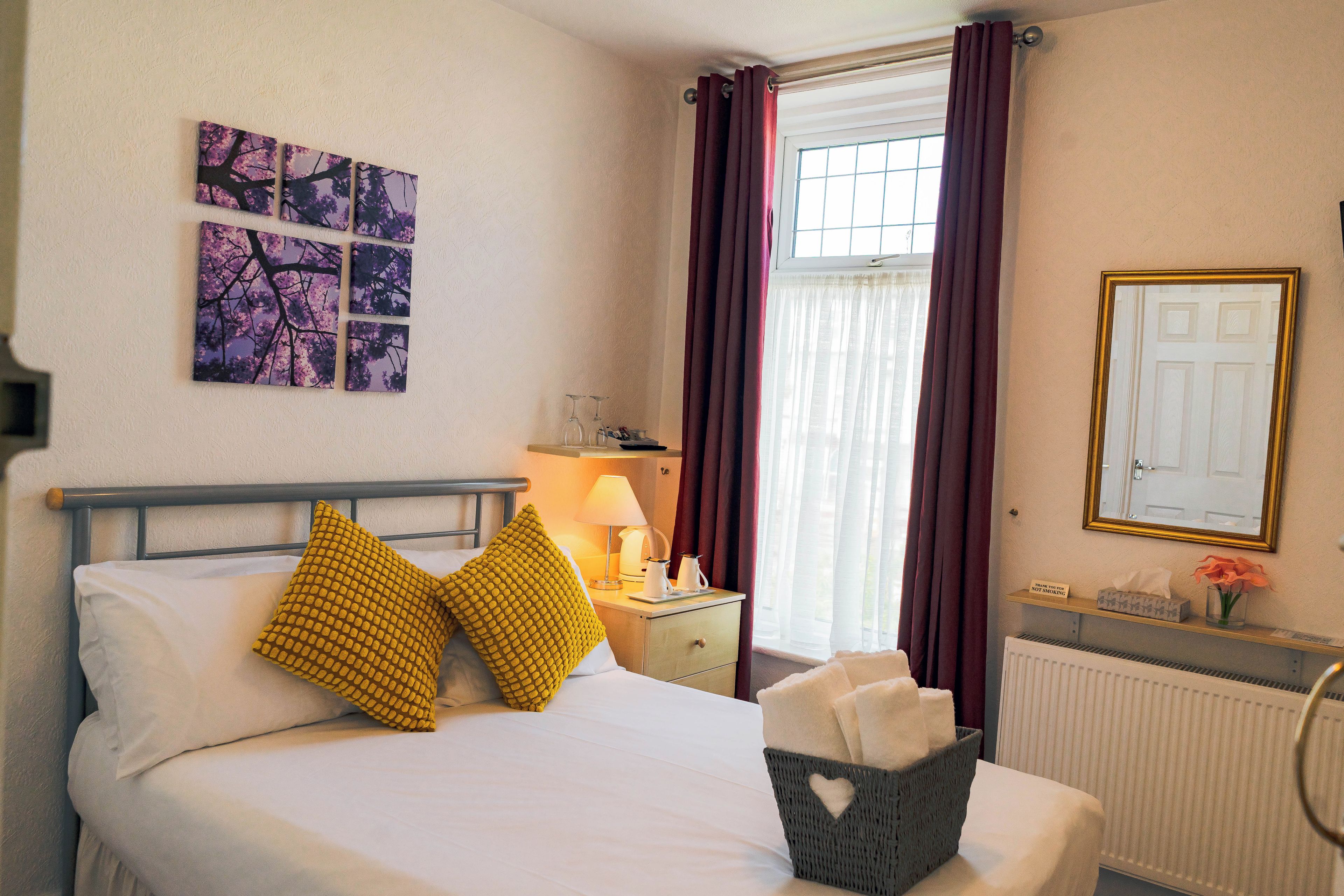 A hotel room with a bed, a nightstand, a lamp, a window with curtains, a radiator, and a wall-mounted mirror.