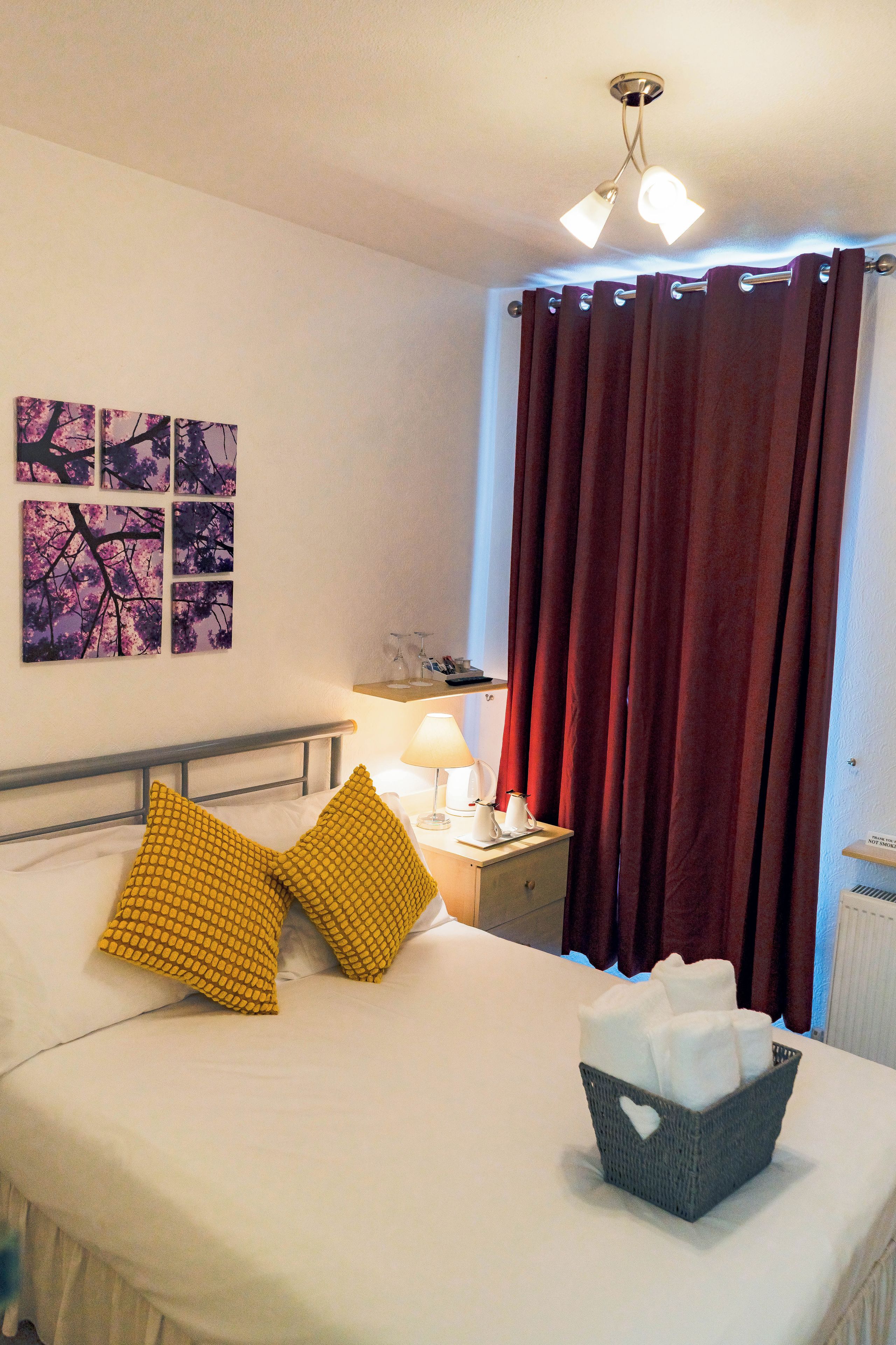 A hotel room with a bed, a nightstand, a lamp, a basket of tissues, and a window with red curtains.